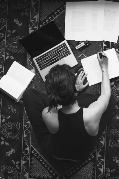 Seen from above, a learner sits on a patterned rug with a laptop, open books and notes